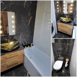 Elegancka łazienka z czarnymi marmurowymi płytkami, złotą umywalką w kształcie łodzi i drewnianą szafką pod umywalką z złotymi uchwytami; biała wanna i toaleta z czarnym panelem sterowania.