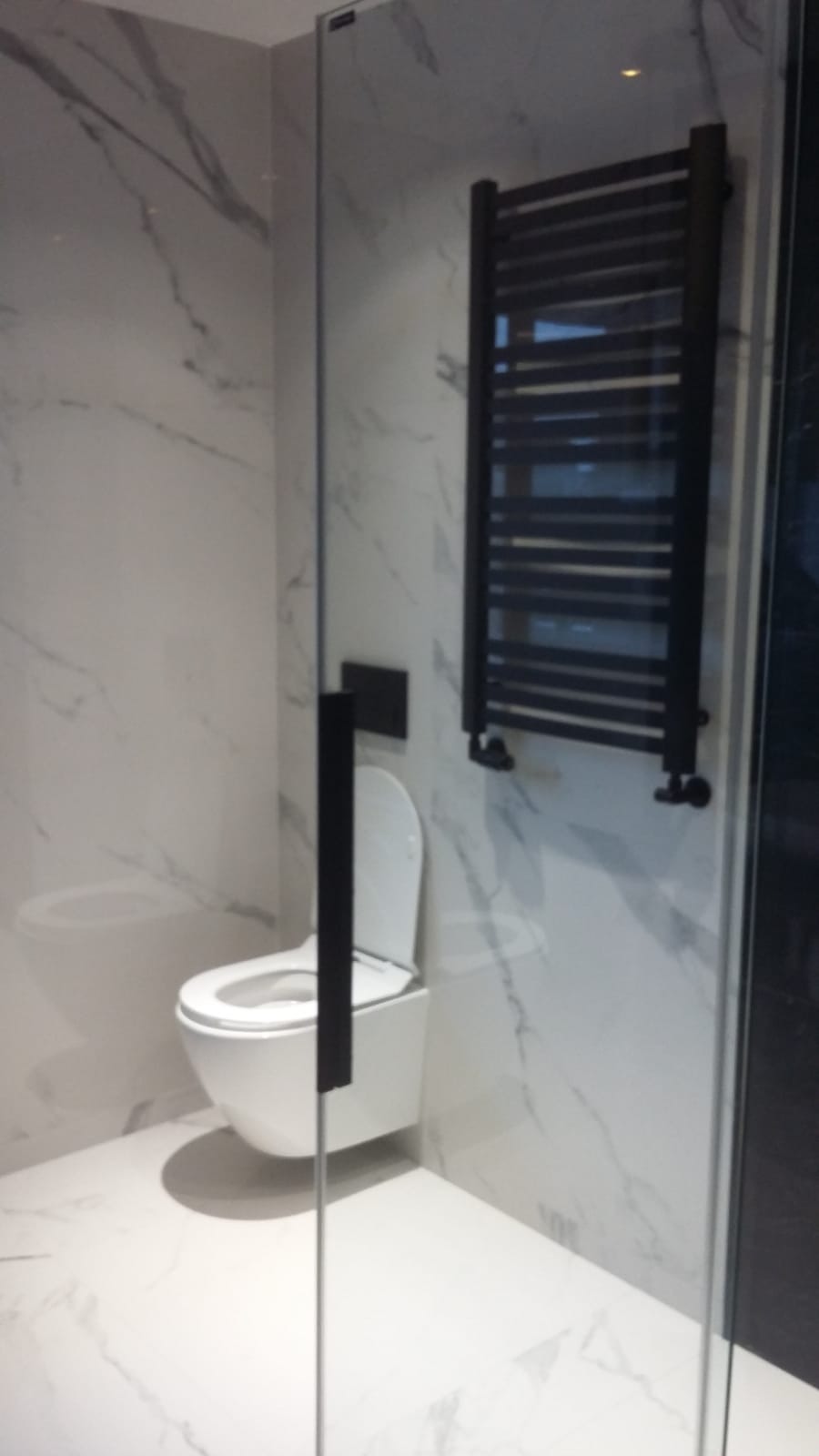 Nowoczesna łazienka z marmurowymi płytkami, wiszącą toaletą i czarnym grzejnikiem drabinkowym, widok przez szklaną kabinę prysznicową.