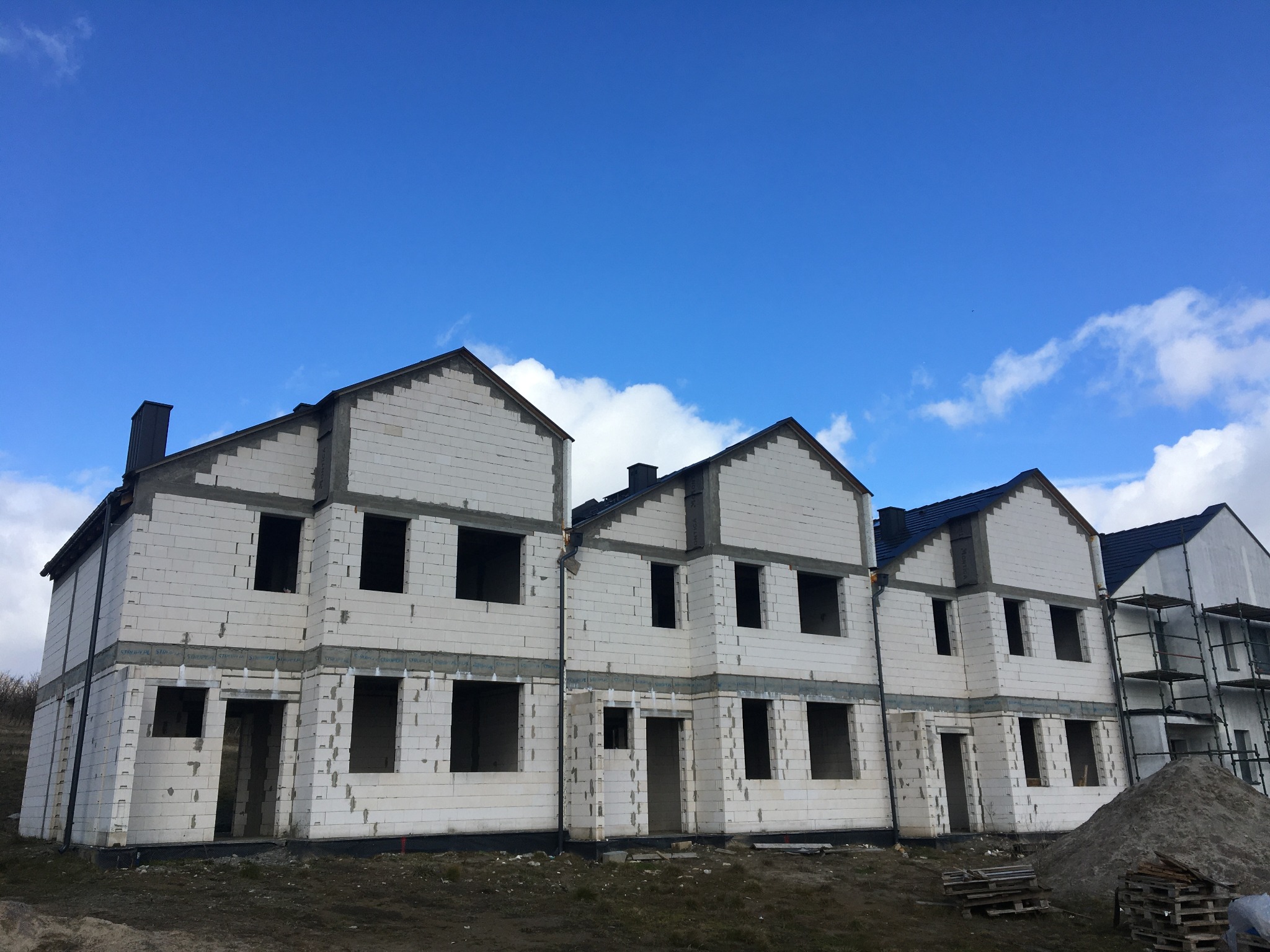 Surowe, dwukondygnacyjne budynki w stanie surowym zamkniętym z białych bloczków, widoczne na tle błękitnego nieba z chmurami, z paletami i stertą piasku na pierwszym planie.