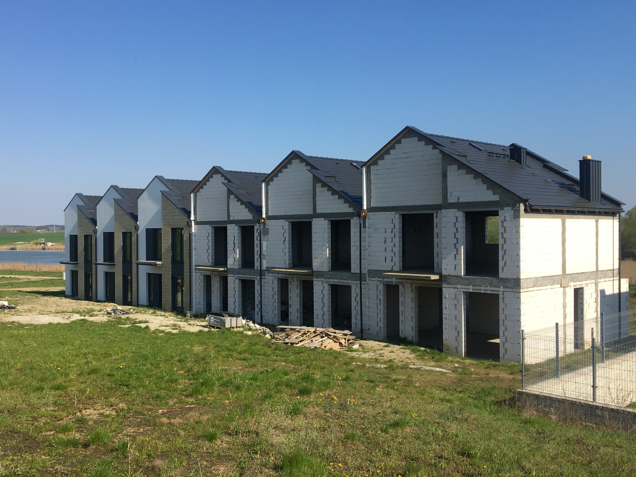 Szeregówka domów w stanie surowym otwartym, z widoczną konstrukcją z bloczków, pokryta ciemną dachówką, na tle zielonej łąki i błękitnego nieba.