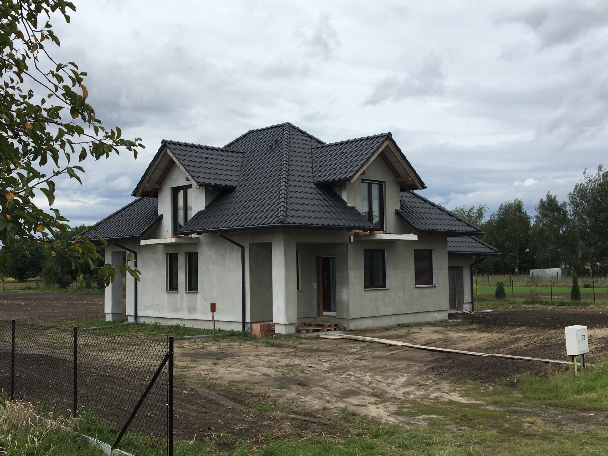 Surowy, dwukondygnacyjny dom z ciemną dachówką, w trakcie budowy, z widocznymi cegłami i paletami przy wejściu, otoczony świeżo zaoraną ziemią i ogrodzeniem z siatki.