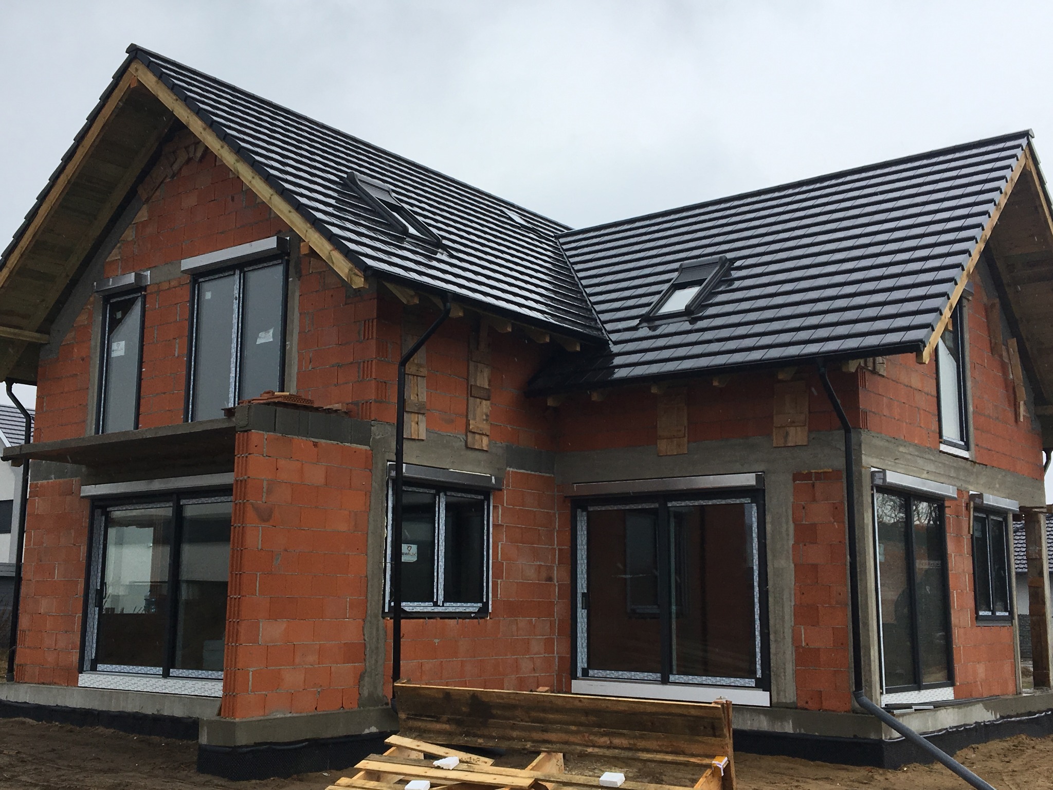 Budowa domu jednorodzinnego w stanie surowym otwartym: ściany z czerwonej cegły, zamontowane okna z roletami zewnętrznymi, czarny dach z dachówką ceramiczną i oknami połaciowymi, widoczne elementy...
