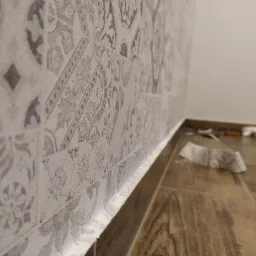 Fragment ściany wykończonej wzorzystą tapetą w odcieniach szarości, z widoczną listwą przypodłogową i fragmentem drewnopodobnej podłogi. W tle widoczne narzędzia i resztki tapety.