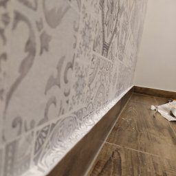 Usługi Ogólnobudowlane Michał Ruszyński - Ściana wyłożona wzorzystymi płytkami ceramicznymi w odcieniach szarości, z widoczną fugą i fragmentem podłogi imitującej drewno.