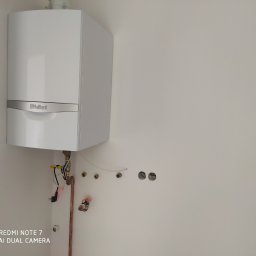 Usługi Ogólnobudowlane Michał Ruszyński - Biały, wiszący piec gazowy marki Vaillant na białej ścianie, z widocznymi rurami miedzianymi i otworami w ścianie przygotowanymi do podłączenia.