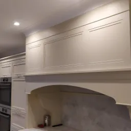 Jasna, klasyczna kuchnia z zabudową meblową, piekarnikiem i okapem nad płytą grzewczą z marmurowym panelem. Oświetlenie punktowe w suficie.