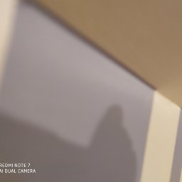 Usługi Ogólnobudowlane Michał Ruszyński - Fragment ściany pomalowanej na szaro z widocznym cieniem, z paskami w rogu pomieszczenia i logo telefonu na dole.