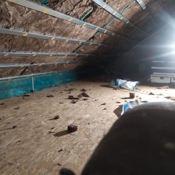 Usługi Ogólnobudowlane Michał Ruszyński - Poddasze w trakcie ocieplania wełną mineralną, widoczne metalowe profile mocujące i porozrzucane fragmenty wełny na podłodze z płyty OSB, oświetlone jasnym światłem.