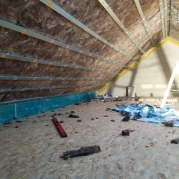 Usługi Ogólnobudowlane Michał Ruszyński - Poddasze w trakcie ocieplania: widoczna wełna mineralna między metalowymi profilami, poziomica, resztki materiału izolacyjnego i folii na podłodze z płyt OSB.