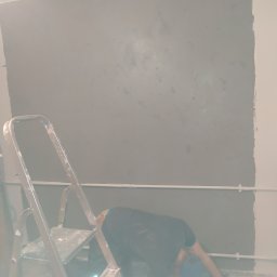 Usługi Ogólnobudowlane Michał Ruszyński - Malowanie ściany na szaro, drabina malarska i osoba w trakcie pracy, widoczne niedomalowane fragmenty w rogu ściany i instalacja elektryczna