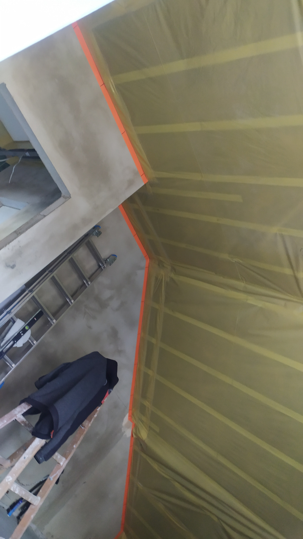 Poddasze w trakcie ocieplania, widoczne krokwie zabezpieczone folią, drabina z kurtką, ściana z oknem po obróbce tynkarskiej, pomarańczowe listwy wyznaczają granice prac.