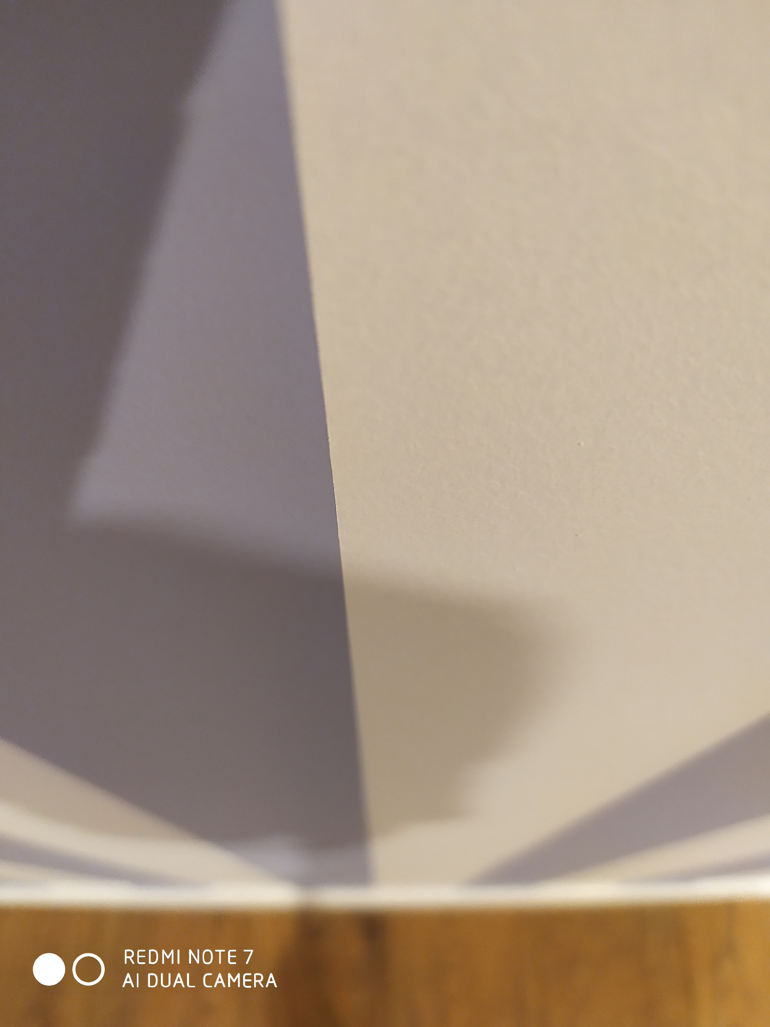 Fragment ściany pomalowanej w pionowe pasy w odcieniach szarości i beżu, widoczne z dołu, z dolną krawędzią ściany i kawałkiem podłogi w tle.