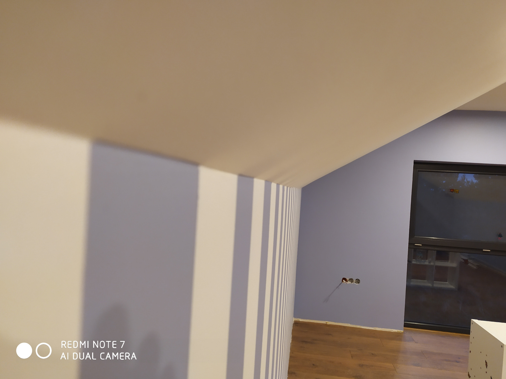 Pomalowane na fioletowo wnętrze pokoju z drewnianą podłogą, widoczne gniazdka elektryczne i okno, ściana z pionowymi białymi pasami.