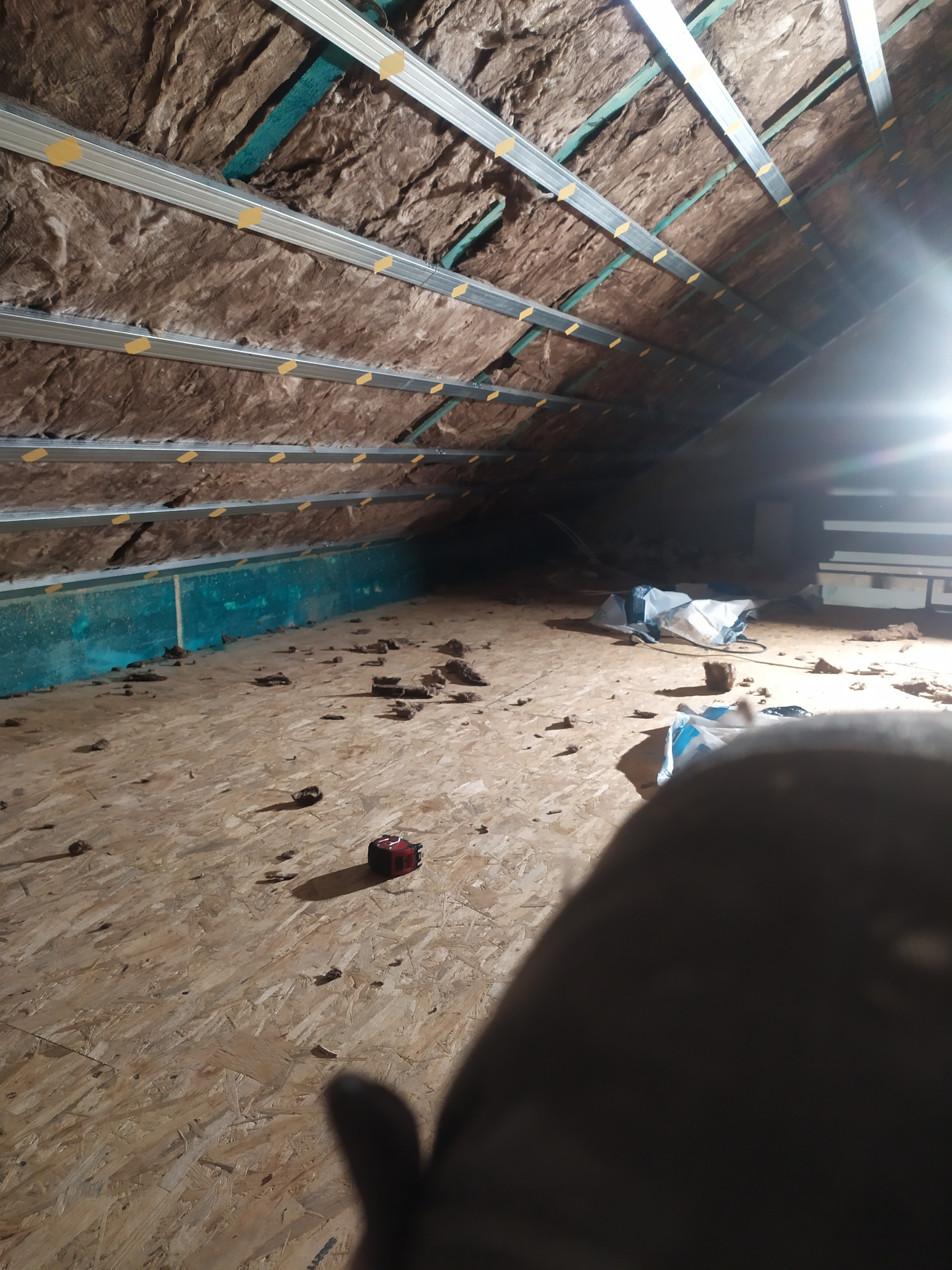 Poddasze w trakcie ocieplania wełną mineralną, widoczne metalowe profile mocujące i porozrzucane fragmenty wełny na podłodze z płyty OSB, oświetlone jasnym światłem.