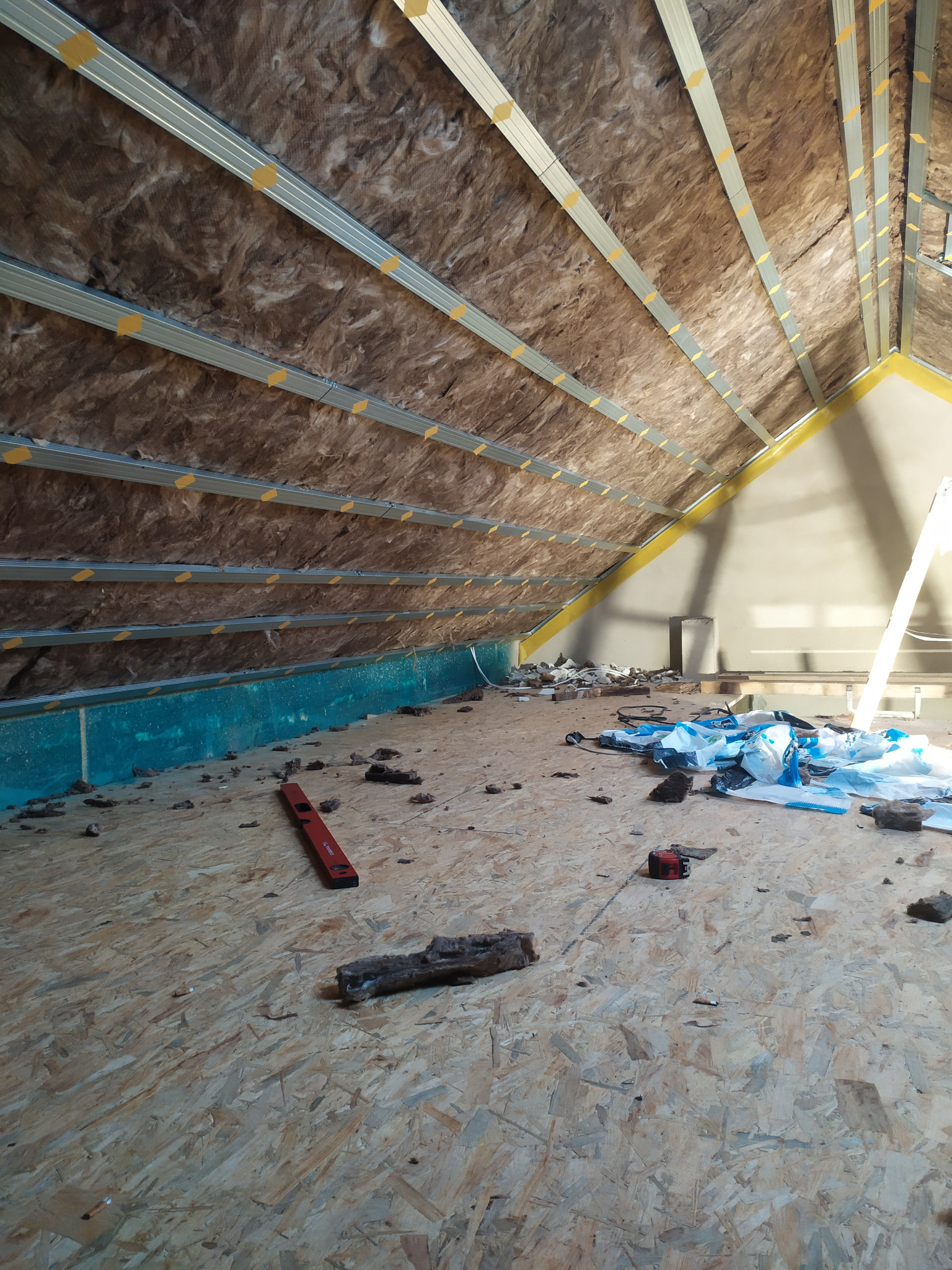 Poddasze w trakcie ocieplania: widoczna wełna mineralna między metalowymi profilami, poziomica, resztki materiału izolacyjnego i folii na podłodze z płyt OSB.