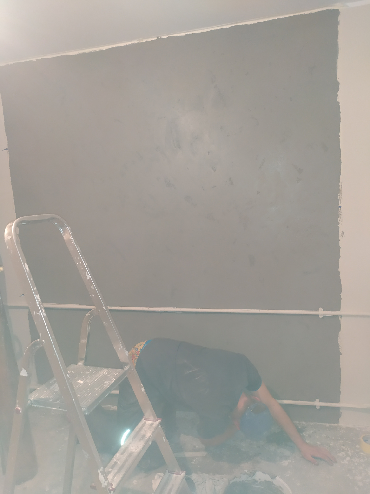 Malowanie ściany na szaro, drabina malarska i osoba w trakcie pracy, widoczne niedomalowane fragmenty w rogu ściany i instalacja elektryczna