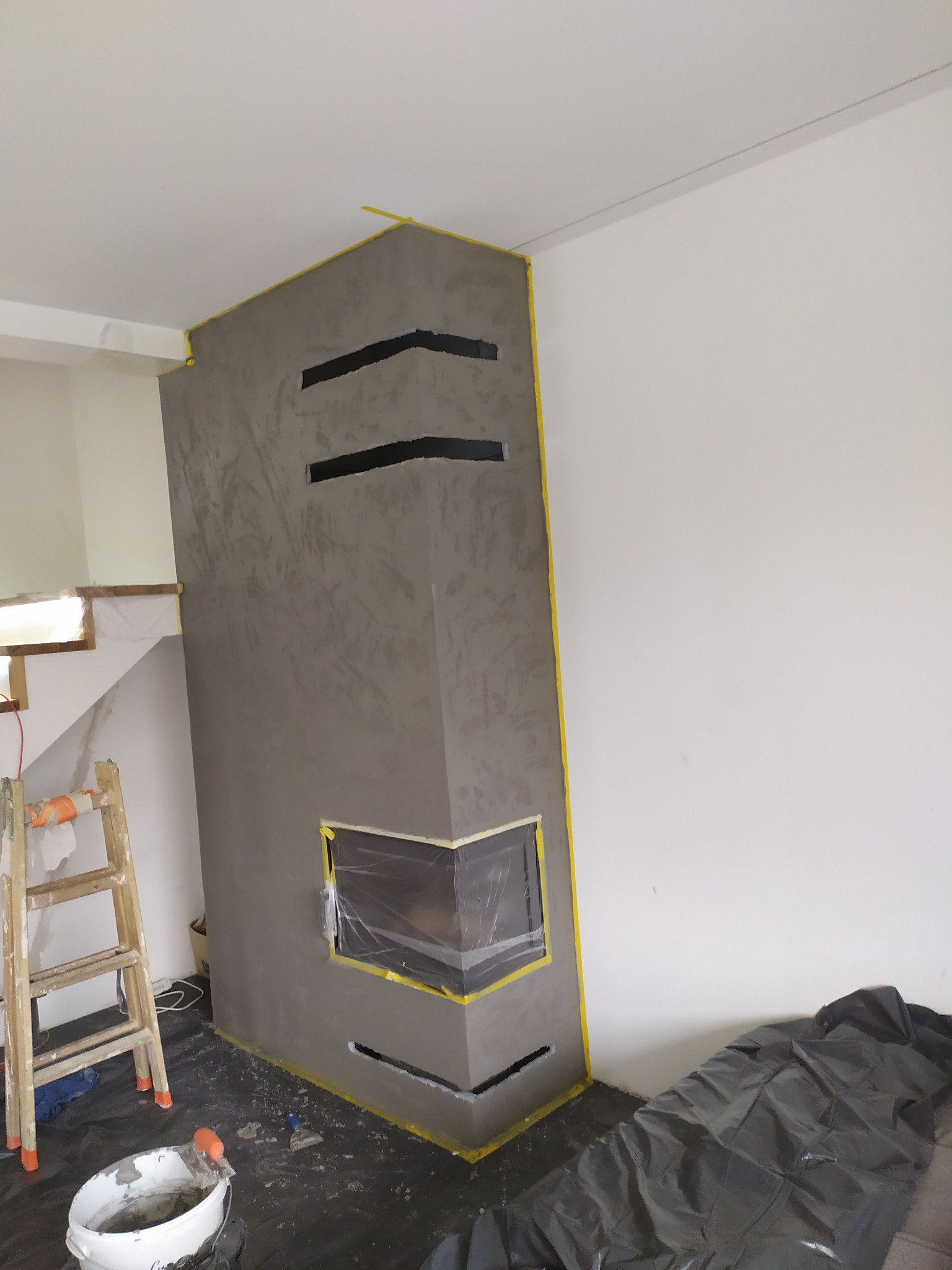 Szary kominek z dekoracyjnym tynkiem, zabezpieczony taśmą malarską, z widocznym wiadrem i drabiną, w trakcie prac wykończeniowych.