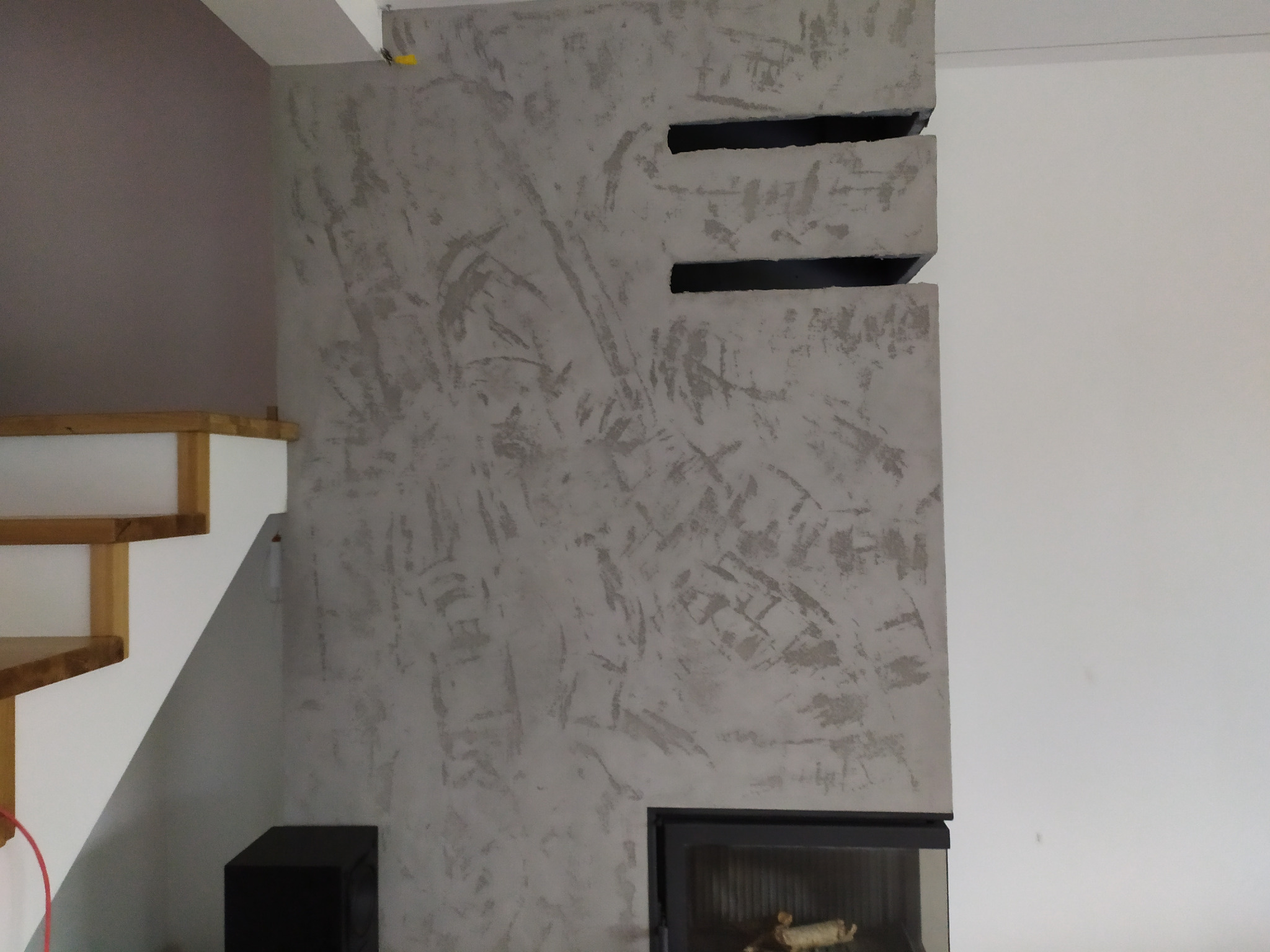 Ściana z dekoracyjnym tynkiem w odcieniach szarości, z trzema poziomymi wgłębieniami w górnej części, obok drewnianych schodów i kominka z szybą.