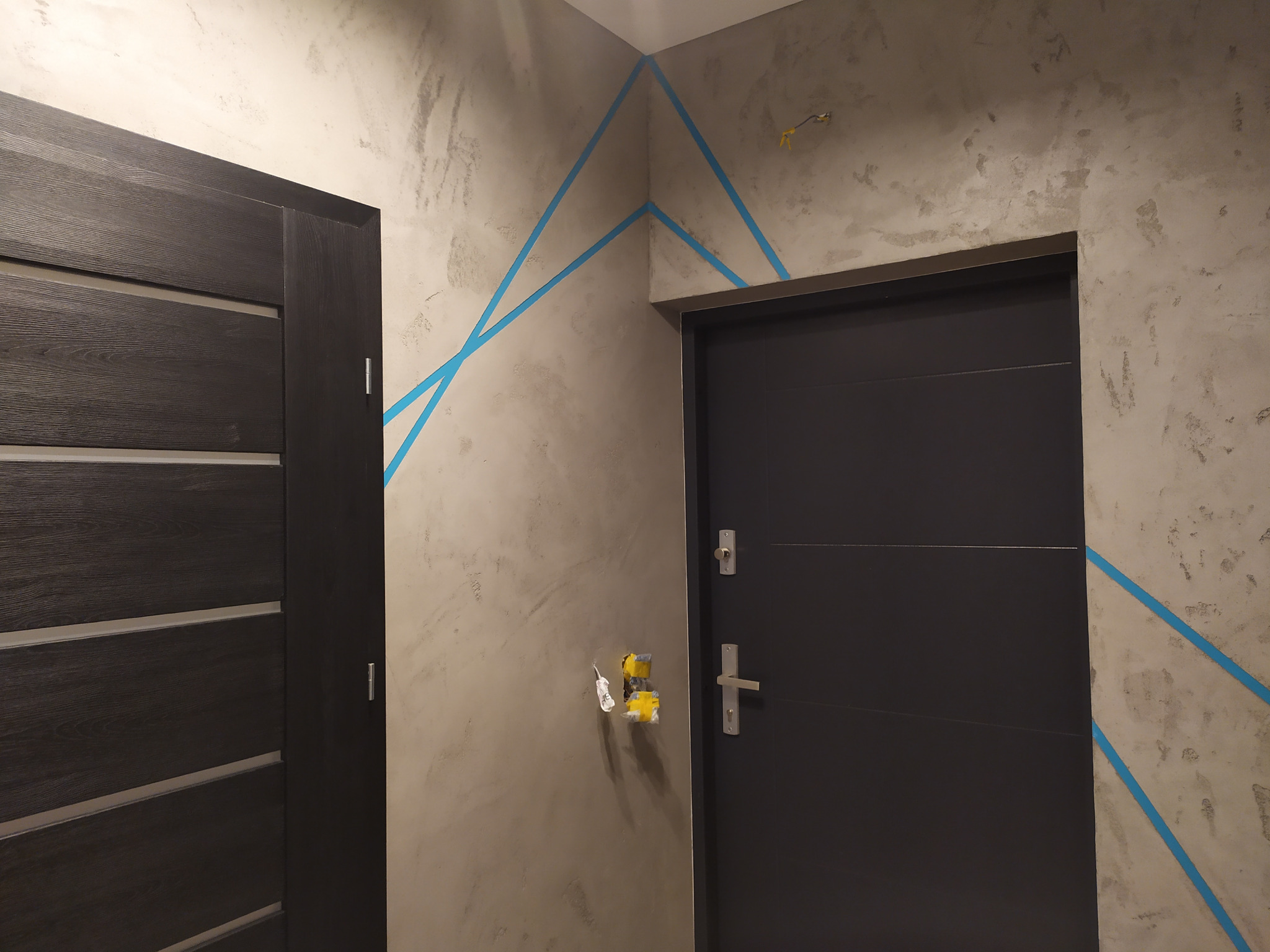 Ściana z efektem dekoracyjnym, zabezpieczona niebieską taśmą malarską, widoczne dwie ciemne, nowoczesne drzwi, gniazdko elektryczne zabezpieczone taśmą.