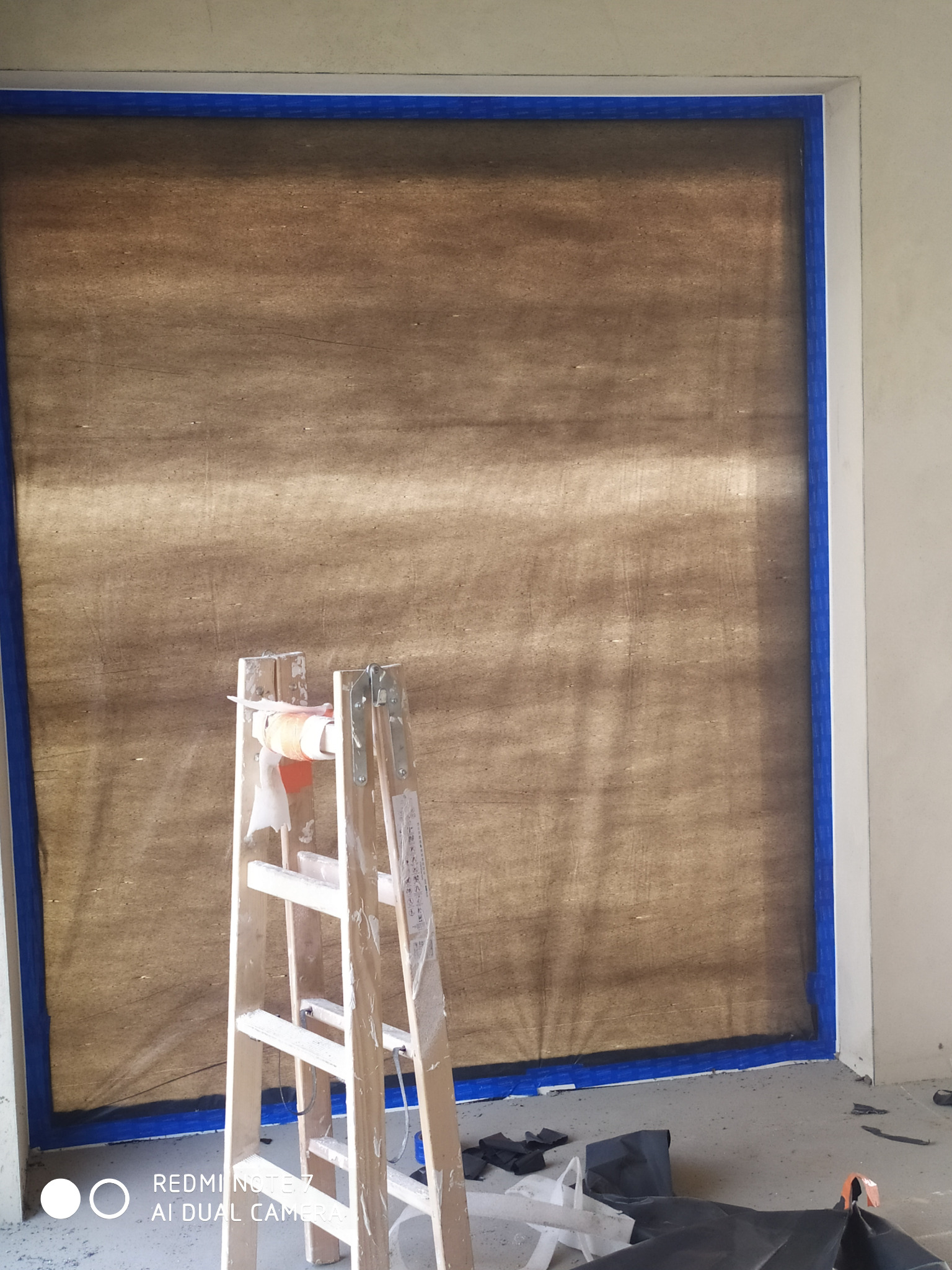 Przygotowanie do prac malarskich: okno zabezpieczone folią i taśmą malarską, drabina malarska, widoczne ślady farby na podłodze.