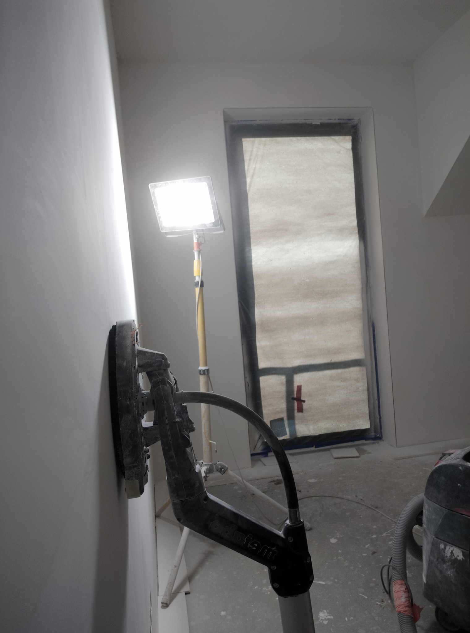 Szlifowanie ściany szlifierką z odkurzaczem w pomieszczeniu przygotowywanym do malowania w miejscowości Linia, widoczne okno zabezpieczone folią i lampa oświetleniowa.