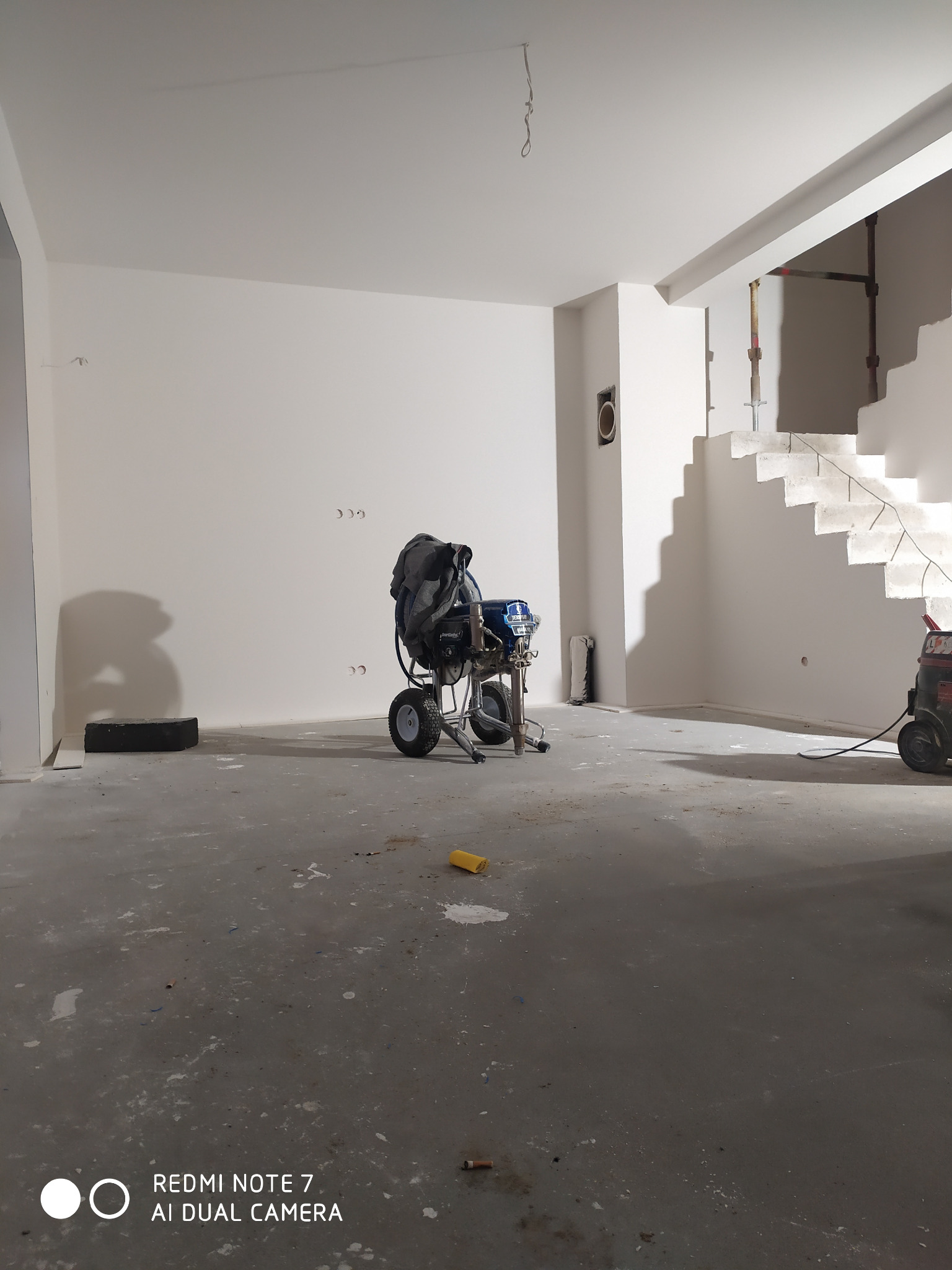 Wnętrze w trakcie odświeżania z maszyną do natryskowego nakładania farby na pierwszym planie, widoczne surowe betonowe schody i ślady farby na podłodze.