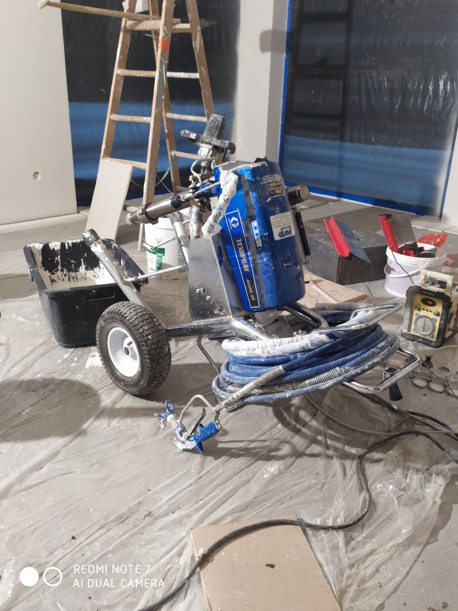 Agregat malarski Texspray Mark VII z wężem i pistoletem natryskowym, stojący na folii ochronnej w pomieszczeniu w trakcie prac malarskich, z widoczną drabiną i zabezpieczoną ścianą.