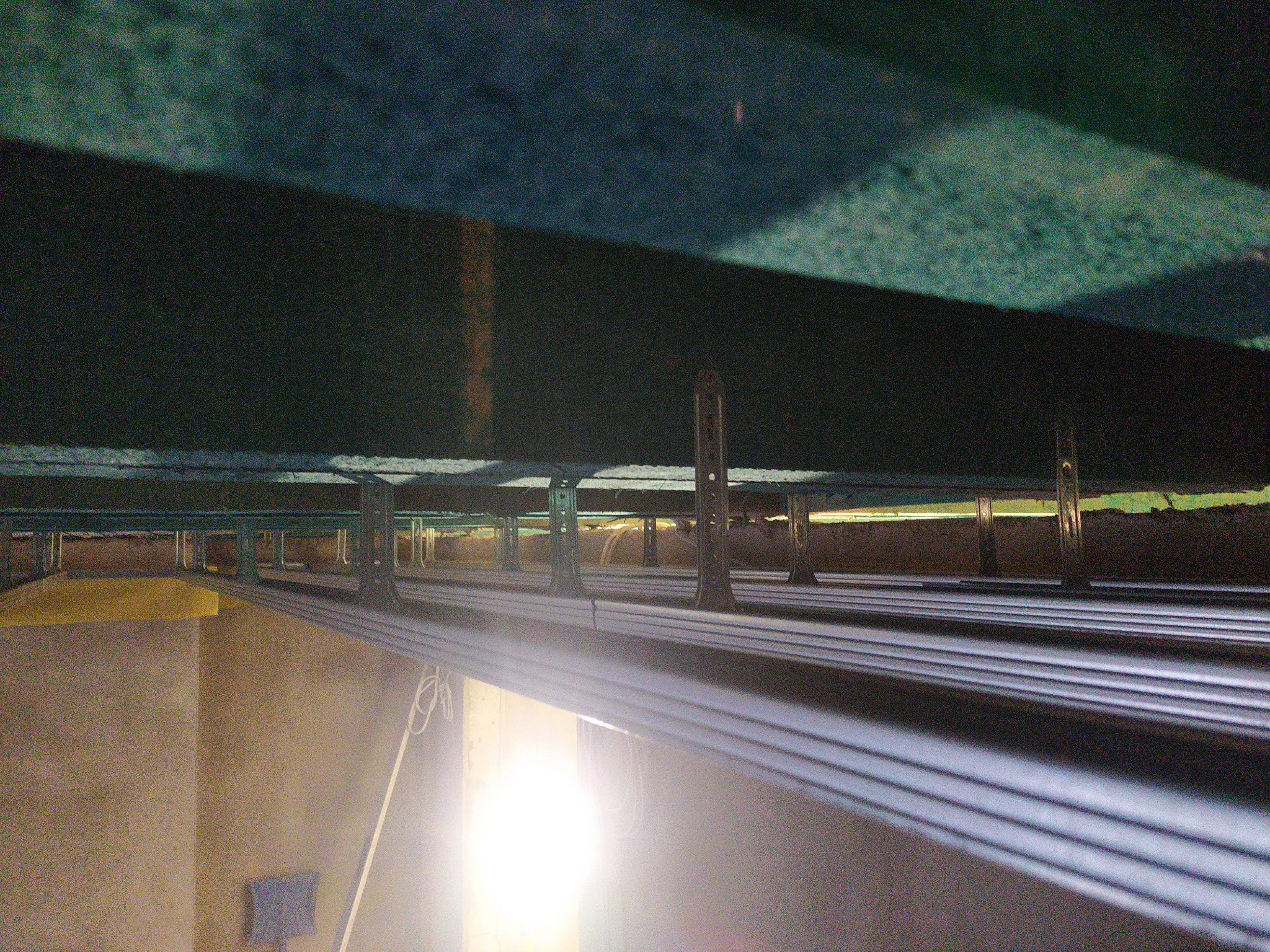 Poddasze w trakcie ocieplania: widoczne metalowe profile mocujące izolację, żółta wełna mineralna oraz konstrukcja dachu. Silne światło w dolnej części kadru.