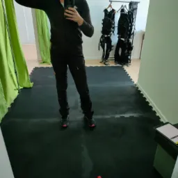 Osoba w sportowym stroju robi selfie w lustrze na tle sali treningowej z matą, hantlami i wieszakiem z ubraniami do treningu EMS.