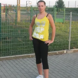 Kobieta w sportowym stroju pozuje na tle ogrodzenia boiska. Żółta koszulka, czarne legginsy 3/4 i białe buty. Widoczny fragment kosza do koszykówki i trawa.