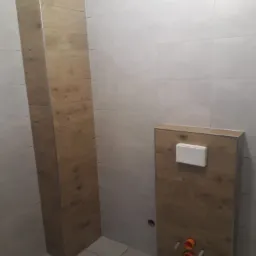 Stelaż podtynkowy WC obudowany panelami imitującymi drewno, widoczne przyłącza wodno-kanalizacyjne i biały przycisk spłukujący, na tle białych płytek ceramicznych.