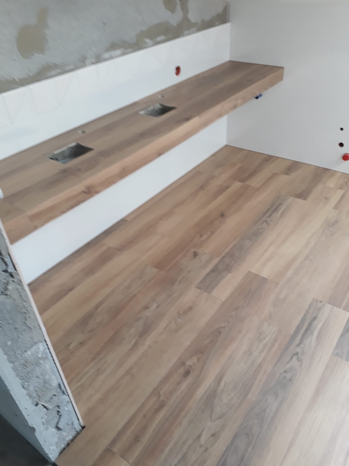 Niedokończona łazienka z drewnopodobną podłogą, drewnianym blatem z wyciętymi otworami na umywalki i częściowo wykończonymi ścianami.