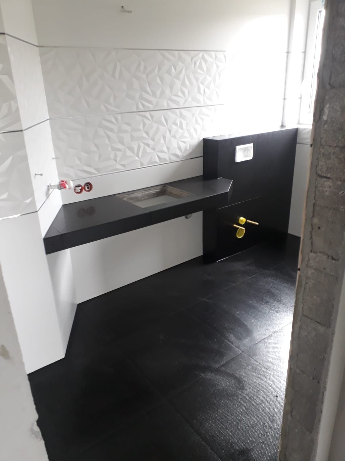 Częściowo wykończona łazienka z czarną podłogą i blatem pod umywalkę, białe ściany z dekoracyjnymi płytkami 3D, widoczne przyłącza hydrauliczne i fragment surowego muru.