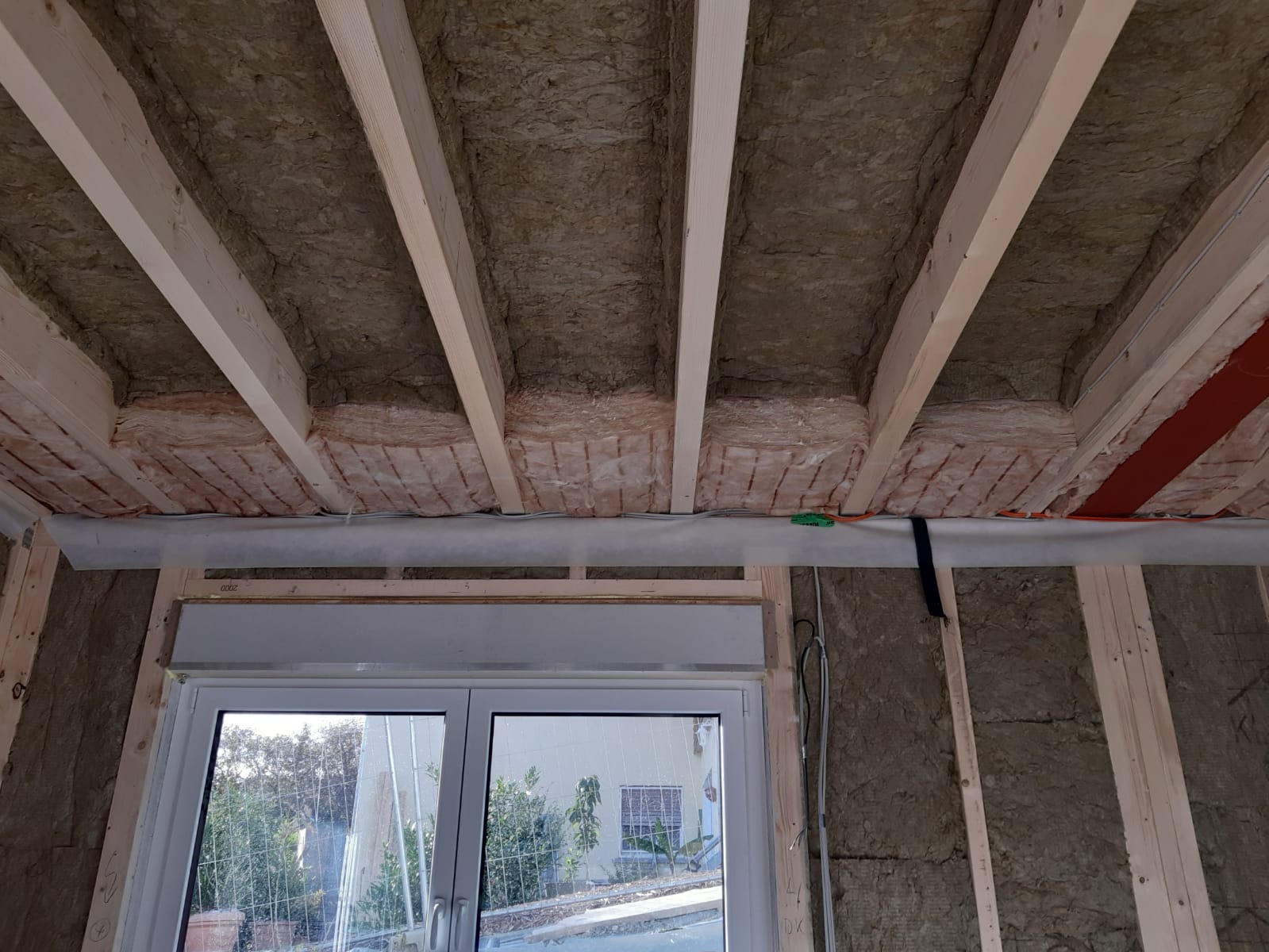 Izolacja ścian i sufitu wełną mineralną w trakcie montażu, widoczne drewniane belki konstrukcyjne i okno z widokiem na zewnątrz.