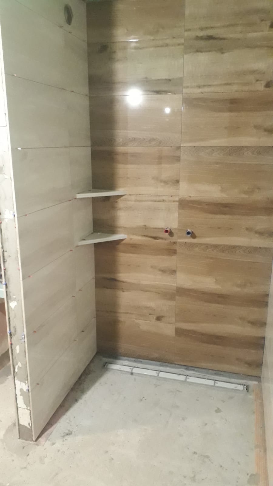 Wnętrze kabiny prysznicowej w trakcie prac wykończeniowych, widoczne płytki imitujące drewno na ścianie, dwie półki w rogu oraz odpływ liniowy w podłodze.