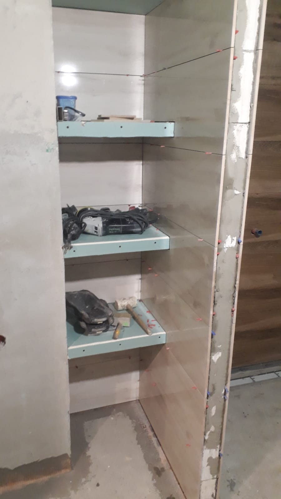 Wnętrze w trakcie tworzenia zabudowy z półkami, widoczne narzędzia i poziomice, ściany przygotowane do położenia płytek.