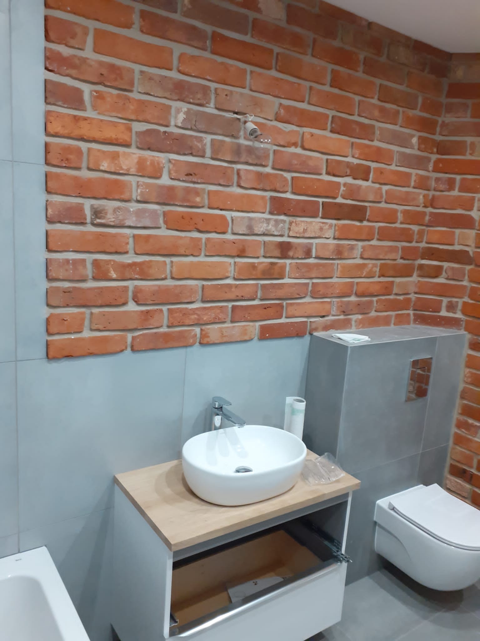 Nowoczesna łazienka z umywalką nablatową, toaletą podwieszaną i ścianą z cegły klinkierowej, oświetlenie punktowe na ścianie.