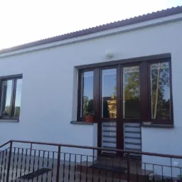 Jasnoszara elewacja budynku jednorodzinnego z brązową stolarką okienną i metalową balustradą, widoczna doniczka z rośliną na parapecie okna.