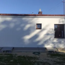 Boczny widok na budynek z białą elewacją, dwoma oknami o brązowych ramach i dwoma małymi okienkami piwnicznymi, cień drzewa na ścianie, zielony wąż ogrodowy leżący na ziemi.