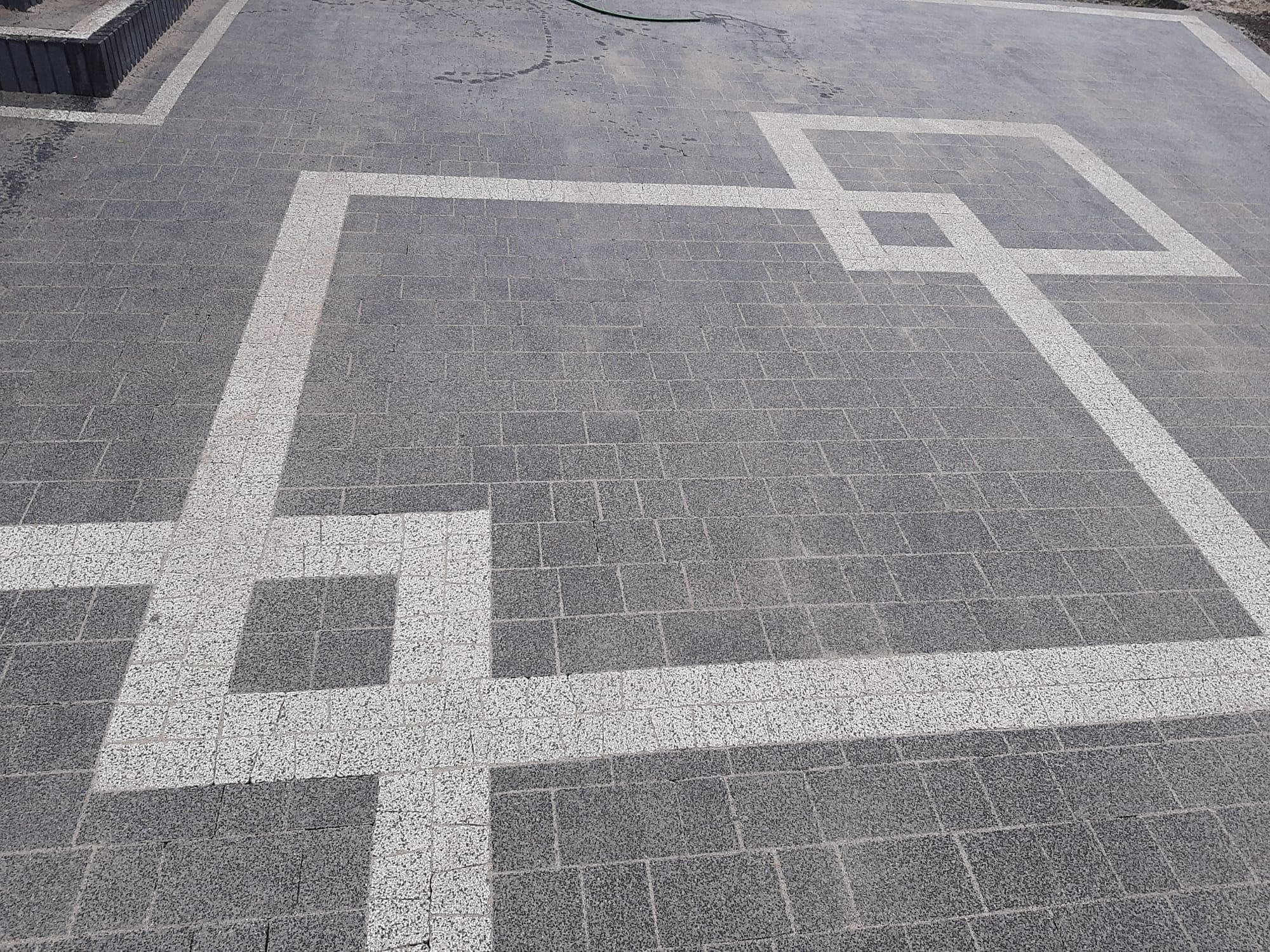 Szara kostka brukowa ułożona we wzór z białymi liniami tworzącymi geometryczny, kwadratowy ornament. Widoczne ślady po wężu ogrodowym.
