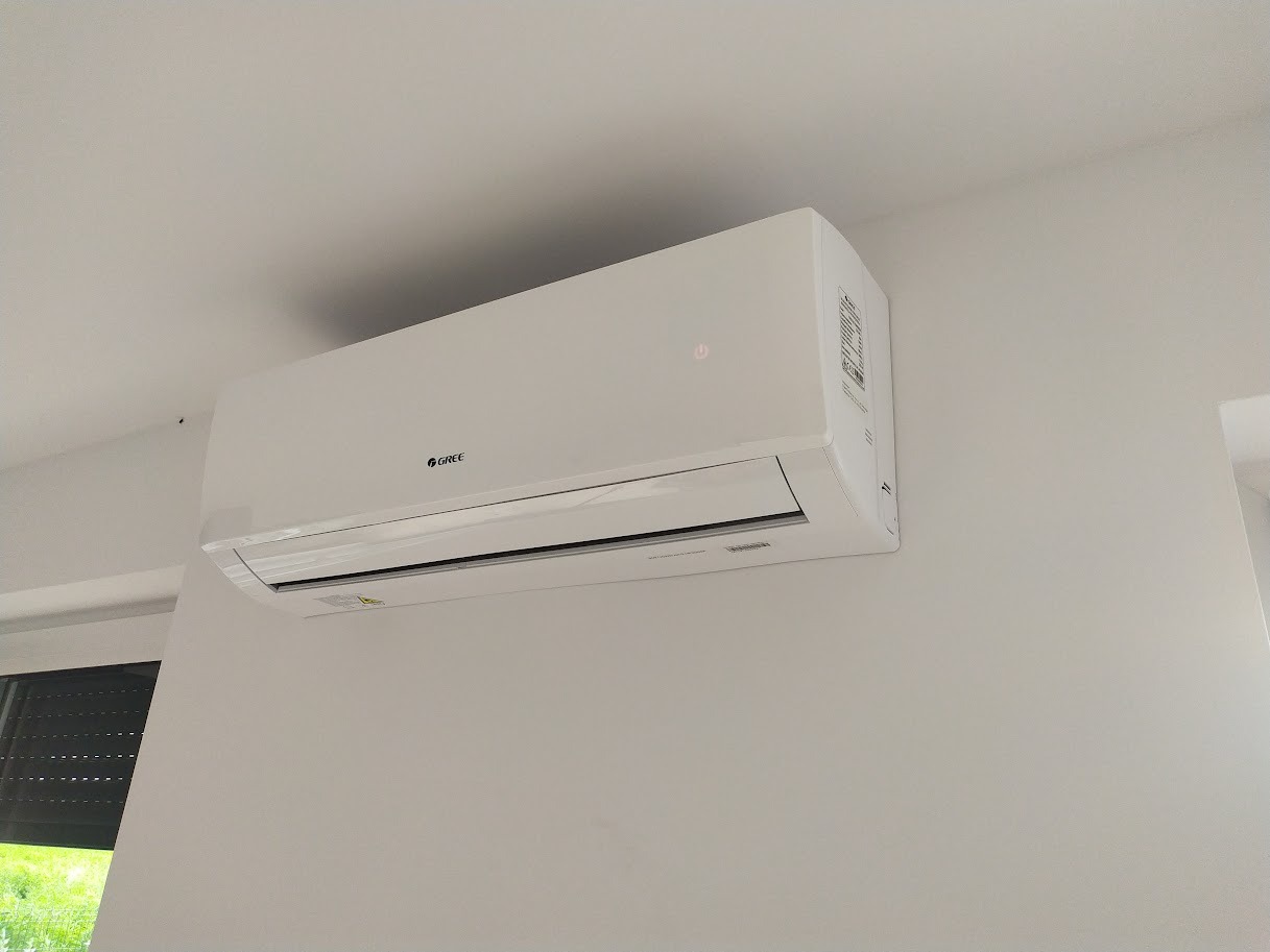 Biała jednostka wewnętrzna klimatyzacji marki Gree, zamontowana na jasnej ścianie w pomieszczeniu z żaluzjami okiennymi.