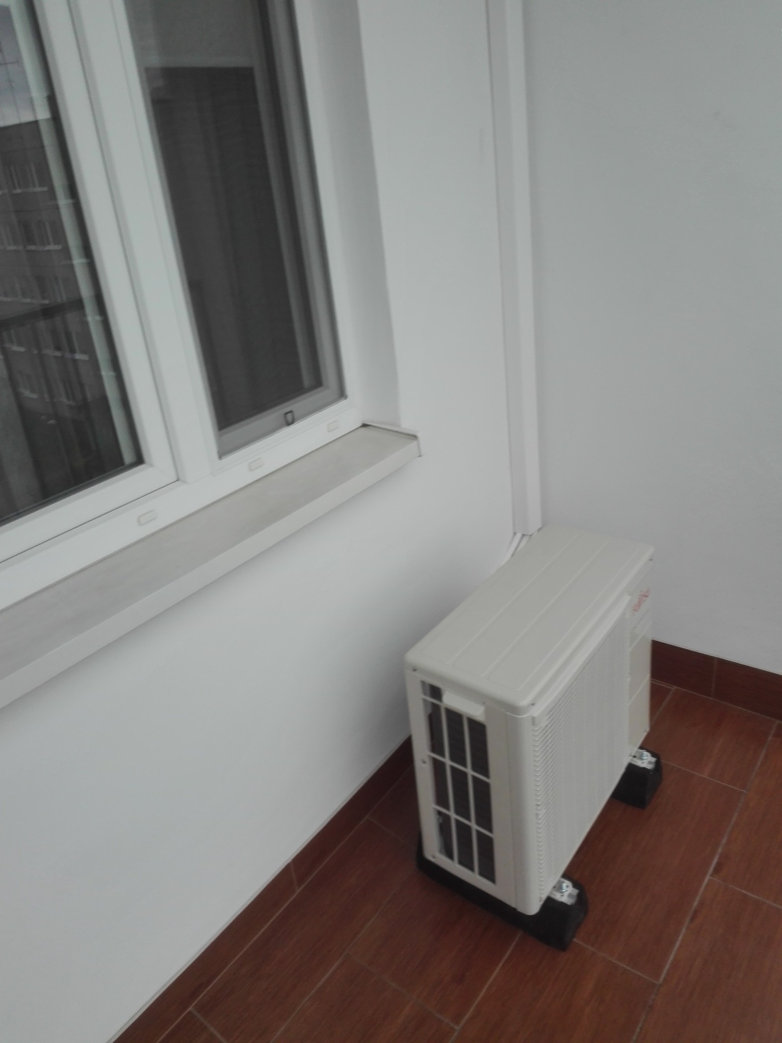 Biała jednostka zewnętrzna klimatyzacji, zamontowana na gumowych podkładkach, stoi na brązowej podłodze z płytek przy białej ścianie i oknie.