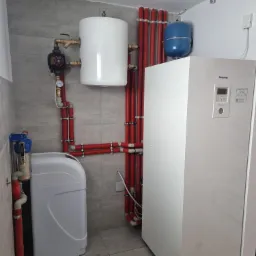 Wnętrze kotłowni z zainstalowaną pompą ciepła Panasonic Aquarea, zbiornikiem buforowym, zasobnikiem CWU i zmiękczaczem wody, połączonymi czerwonymi rurami w izolacji.