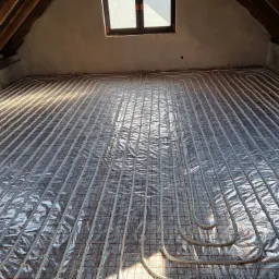 Układ rur ogrzewania podłogowego, widok z góry na rozłożone rury na folii odblaskowej i siatce w trakcie montażu, poddasze z drewnianą konstrukcją więźby dachowej.