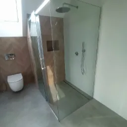 Nowoczesna łazienka z kabiną prysznicową typu walk-in, wykończona płytkami imitującymi drewno i szarą podłogą. Widoczny biały sedes podwieszany oraz deszczownica i bateria prysznicowa.