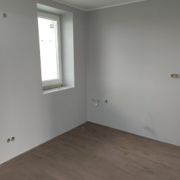 Andrzej Włodarczyk - Pokój po odświeżeniu: szare ściany, brązowe panele podłogowe imitujące drewno, widoczne gniazdka elektryczne przed montażem osprzętu, okno z uszczelnieniem pianką.