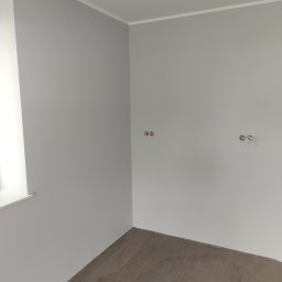 Andrzej Włodarczyk - Pomalowane na szaro wnętrze z brązową podłogą z płytek imitujących drewno, widoczne puszki elektryczne na ścianie i biała listwa przypodłogowa.