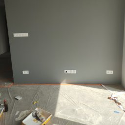 Andrzej Włodarczyk - Świeżo pomalowana szara ściana z widocznymi gniazdkami elektrycznymi i włącznikiem światła, zabezpieczona folią malarską na podłodze, widoczne narzędzia malarskie w kuwecie.