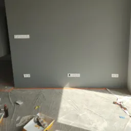 Świeżo pomalowana szara ściana z widocznymi gniazdkami elektrycznymi i włącznikiem światła, zabezpieczona folią malarską na podłodze, widoczne narzędzia malarskie w kuwecie.