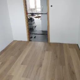 Fragment pokoju z ułożonymi panelami podłogowymi w odcieniach beżu i brązu, widok przez ościeżnicę z widocznym odkurzaczem przemysłowym i elementami remontu w tle.